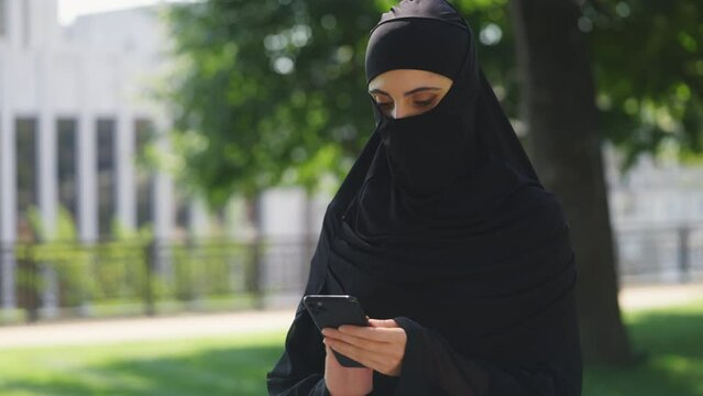 Modern young woman in traditional Muslim burqa using mobile app in city street
