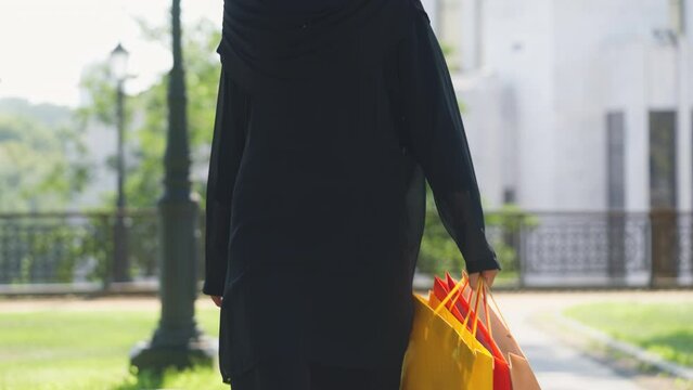 Young woman in black burqa walking city street with colorful shopping bags