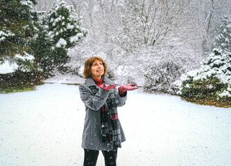 Older woman enjoying falling snow outdoors and catching snowflakes with gloved hands 