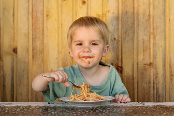 A little happy boy is eating spaghetti. His face is smeared with ketchup
