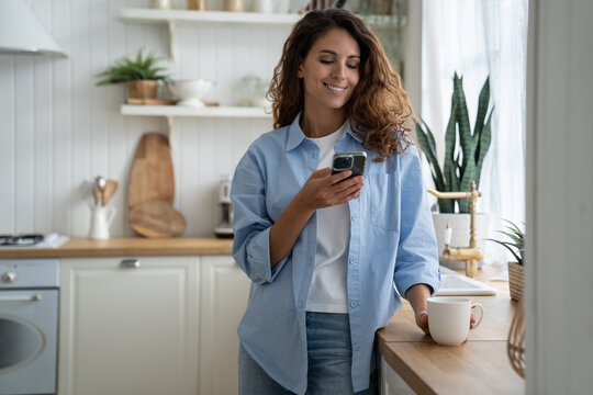Happy woman housewife standing in kitchen with phone chatting with friends in online messenger, drinking coffee. Joyful Italian female in casual shirt flipping through social media feed in smartphone 