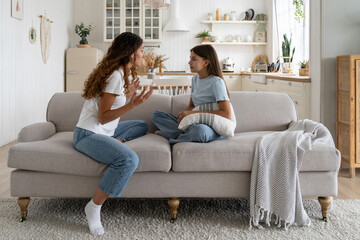 Tense teen girl holding pillow and looking at nervously gesturing mother sits on couch. Annoyed...