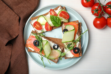Tasty rye crispbreads with salmon, cream cheese and vegetables served on white wooden table, flat lay
