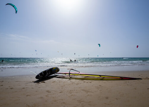 Beach on the coast of light with turquoise blue sea. Board with its sail waiting for windsurfing. In the background unrecognizable kite surfing.