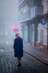 A blonde woman in an overcoat stands on the sidewalk in the morning fog.