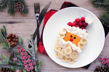 Fun Christmas Santa Claus pancake on a white plate with raspberry hat and banana beard. Above view table scene against a wood background. - obrazy, fototapety, plakaty