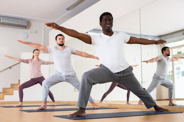 Fototapeta na wymiar Sporty adult African American doing power yoga with group in fitness studio, standing in lunging asana Virabhadrasana..