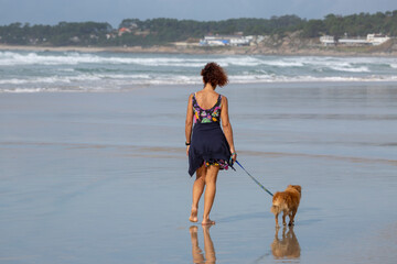 A woman in a flowery dress walks her dog on the beach in summer. A Lanzada Beach, Galicia