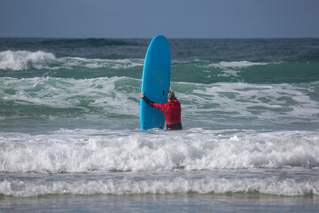 A surfer enters the water at A Lanzada beach with a blue apprentice surfboard