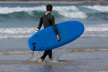 A surfer enters the water at A Lanzada beach with a blue apprentice surfboard