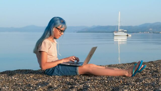 Girl video chat from shore. A teen takes off her headphones and stop computer working to admire the sea with boart under the sun.