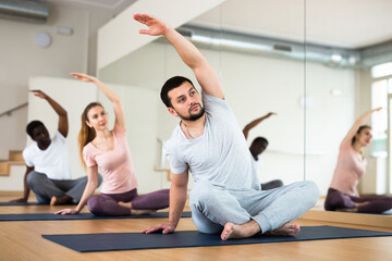 Fototapeta na wymiar Man doing exercises in lotus pose with group, yoga training in gym.