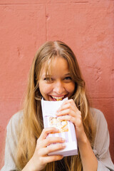 Young woman alone standing against a red wall eating pop corn out of a bag.