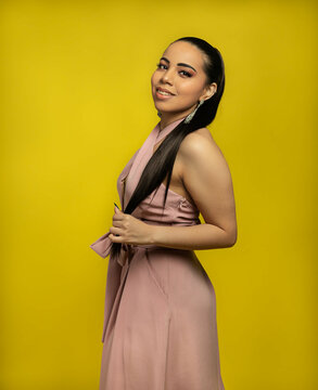 Fashion portrait of woman in pink jumper and yellow backdrop holding long dark hair smiling. 