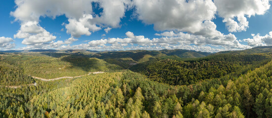 Obraz premium Panoramic aerial photograph of the Salazar valley in Navarre, Spain. Mountains and extensive forests under a blue sky with white clouds. In the center, the village of Ezcároz and a mountain road.