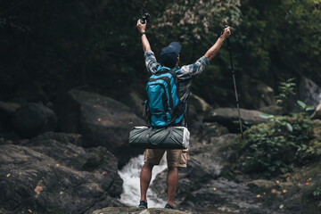 Fototapeta premium Hikers standing on the rock and raising hands to happy with backpacks and background waterfall in the forest. hiking and adventure concept.