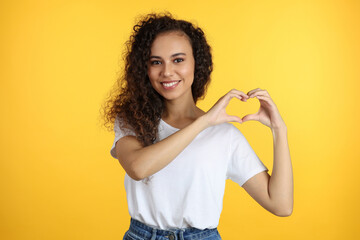 Happy young African-American woman making heart with hands on yellow background