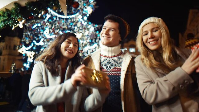 Three happy friends dance with gifts for New Year and Christmas on new year's eve to the sound of chimes in the middle of the street in the lights of the Christmas market against beautiful big