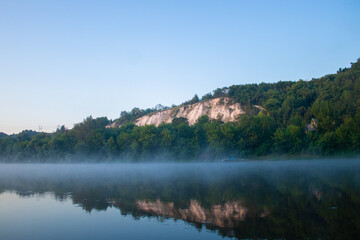 chalk mountain overgrown with dense vegetation, at the foot of the river in the morning haze and a boat near the shore