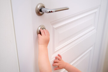 Toddler baby opens the lock, child s hand close-up. White wooden door, metal door handle and baby hand - obrazy, fototapety, plakaty