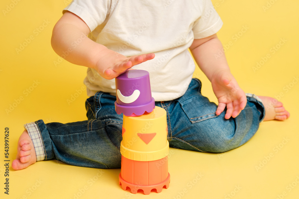 Wall mural Toddler baby is playing logical educational games with a sandbox mold on a studio yellow background. Happy child playing with an educational toy bucket, learning logic. Kid aged one year four months