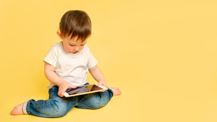 Toddler baby with a digital tablet on a studio yellow background. Happy child playing with a tablet...