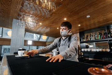A masked barista exquisitely serves ready-made coffee in a modern cafe during a pandemic. Serving ready-made coffee to a client in a cafe.
