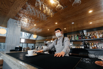 A masked barista exquisitely serves ready-made coffee in a modern cafe during a pandemic. Serving ready-made coffee to a client in a cafe.