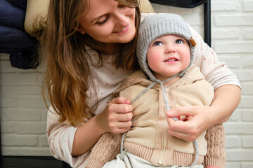 The mother puts a warm hat on the head toddler baby sitting in the home hallway. Woman mom dressing warm clothes on child for winter walk in cold weather. Kid aged one year and three months