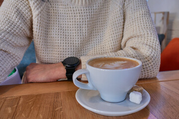 There is a cup of coffee on the table, two pieces of sugar in a saucer. A girl in a white knitted sweater sits at a wooden table in a cafe, she has a smart watch on her hand.