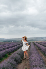 A young beautiful girl in a delicate dress and hat walks through a beautiful field of lavender and enjoys the fragrance of flowers. Rest and beautiful nature. Lavender blooming and flower picking.