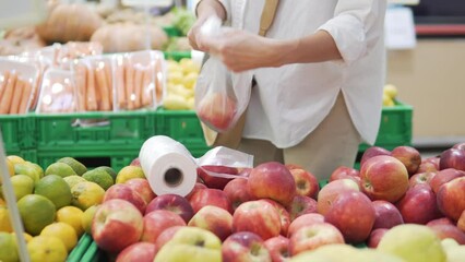 A woman buys red apples at a grocery store. Close-up, women's hands stuff juicy apples into a plastic bag at the store.