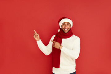 Fototapeta na wymiar Smiling Man Pointing Hand. Positive Multiracial Guy in Santa Hat Pointing Away Paying Your Attention at Empty Space for Advertisement. Indoor Studio Shot Isolated on Red Background 