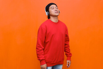 Fototapeta premium Asian man in wireless headphone closes eyes and enjoy song while standing against orange background