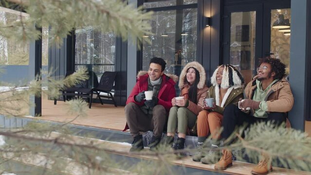 Modern ethnically diverse young friends enjoying spending winter day together sitting on country house porch with cups of hot drinks