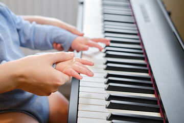 A mother woman teaches a toddler baby to play the piano in a home living room. An adult teacher with a child playing music on an electric piano. Kid boy age one year four months