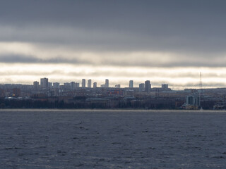 View of the city of Petrozavodsk with silhouettes of high-rise buildings - the capital of the Republic of Karelia on a cloudy day from Lake Onega. The sun's rays partially illuminate the city.