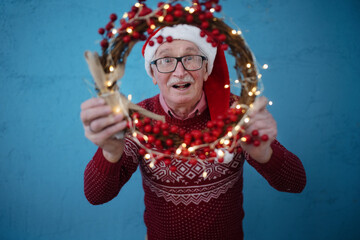 Portrait of happy senior man with Santa Claus hats in studio, looking trough Christmas wreath.