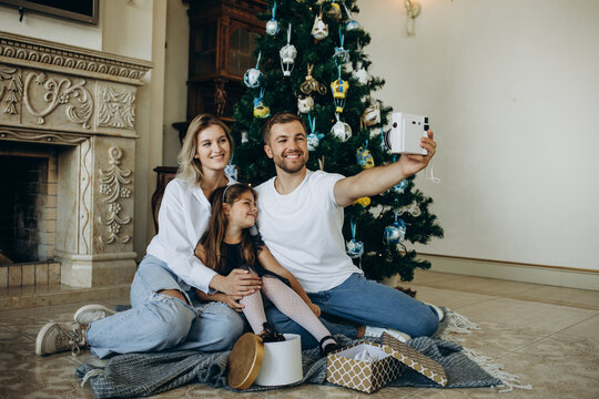 Young happy family of three taking a photo of themselves in living room on Christmas eve. Caucasian family with dog sitting by christmas tree at home taking selfie with mobile phone.