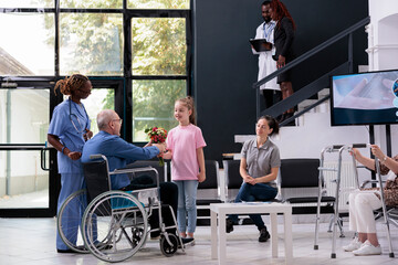 Granddaughter handing bouquet of flowers to disabled grandfather after finishing medical consultation in hospital waiting area. Elderly man discussing health care treatment with nurse