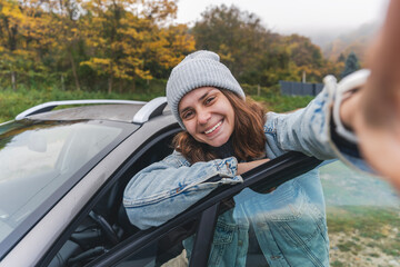 Young cheerful woman driver in a hat and jacket traveling by car taking a selfie on a smartphone...
