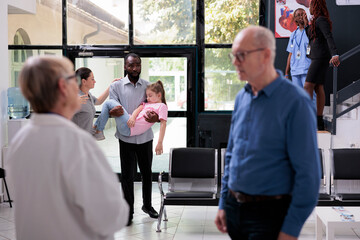 Hospital waiting area full of medical staff and patients ready for medical consultation during checkup visit. Worried family holding fainted unconscious daughter in arms. Medicine concept