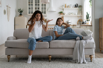 Dumbfounded annoyed angry woman answering call to colleagues at work expressing dissatisfaction with worry after hours. Distant teenage girl sits on couch in living room watching mother swear on phone