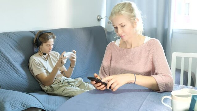 Mother and son sitting at kitchen with smartphones during breakfast and talking