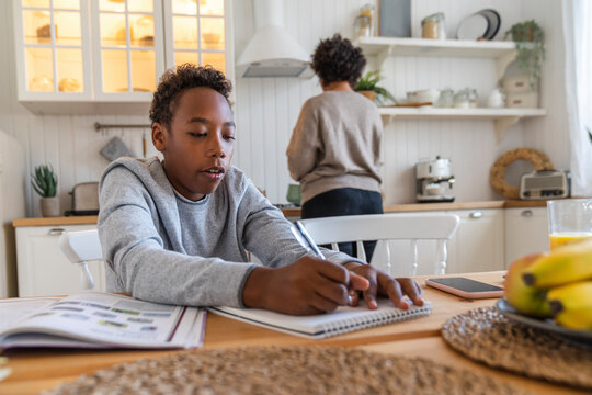 Boy writing on book with mother in background working at home