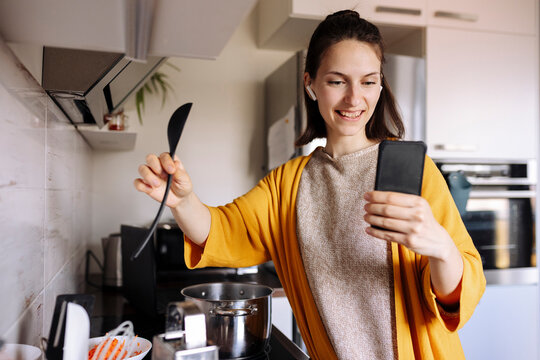 Smiling young woman holding wooden spoon having video call through mobile phone in kitchen