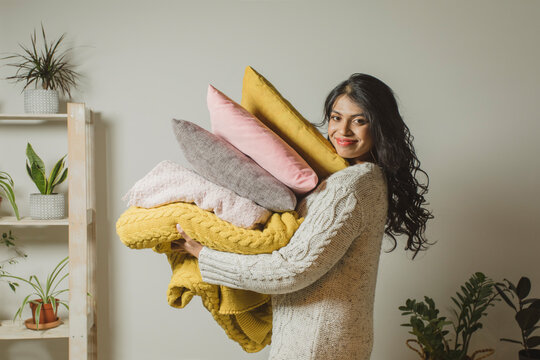 Smiling young woman holding pillows and blankets by wall at home