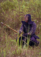 Full-length portrait of kendoka man sitting  in the forest holding the sinai-sword.  Kendo is the Japanese martial art of sword fighting