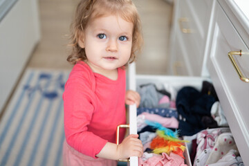 toddler girl arranges clothes into drawer at home - obrazy, fototapety, plakaty