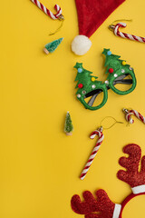 Colourful funny Christmas eyeglasses, candy canes, deer horns, Santa hat, toy fir trees on yellow background. New Year, Christmas accessories for winter holidays party celebration
